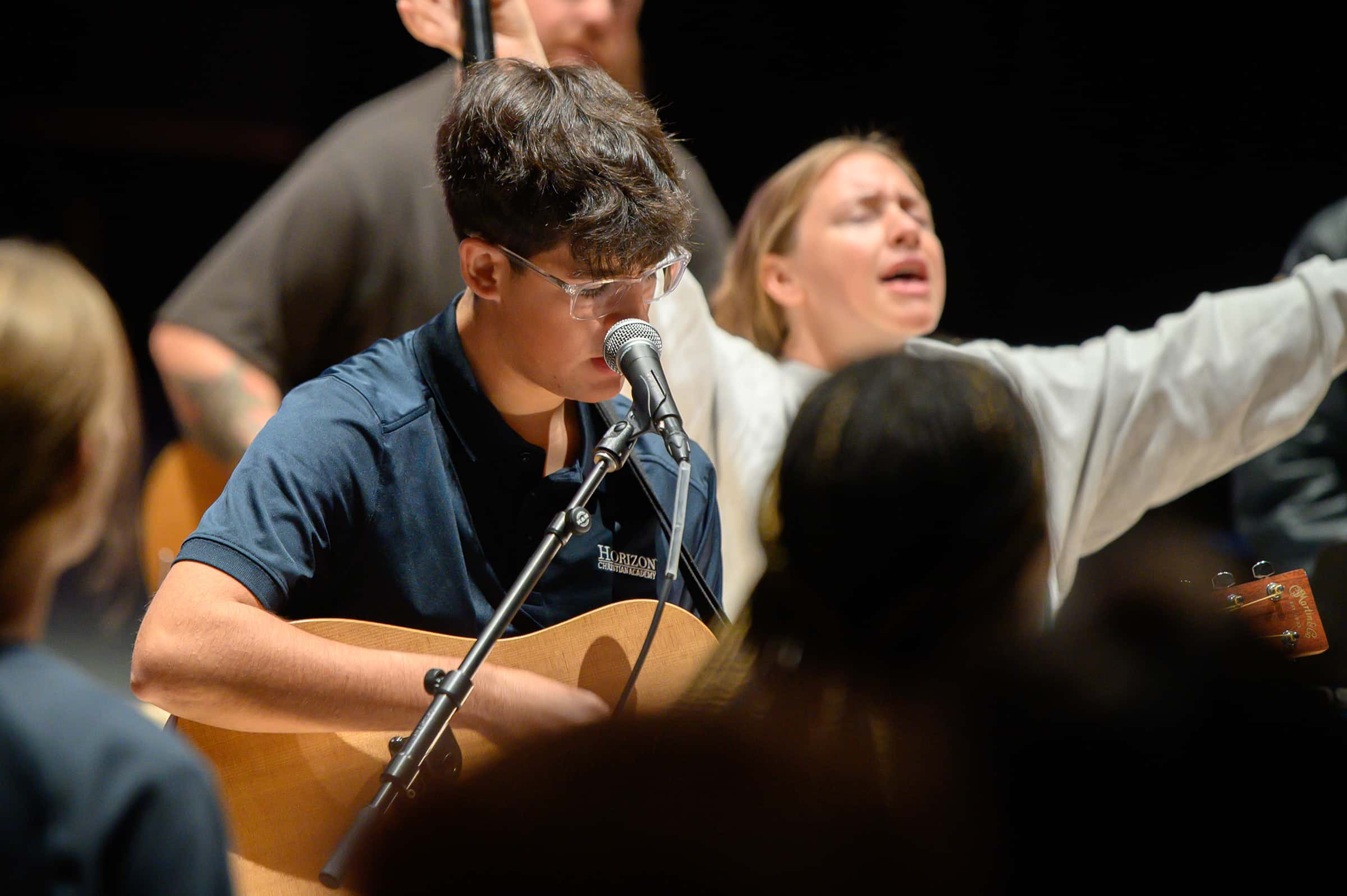 male with glasses playing guitar sining into a microphone